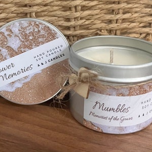 May include: Two silver candle tins with a beach-themed design. The lid of one tin is off, revealing a white soy candle inside. The label on the tin reads "Gower Memories" and "Mumbles Memories of the Gower". Both labels have a hand-poured soy candle logo.