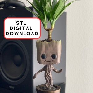 Puede incluir: Maceta impresa en 3D de Groot con una planta verde que crece en la parte superior. La maceta es marrón y tiene una cara sonriente. El texto "STL DIGITAL DOWNLOAD" está en un cuadro rojo.