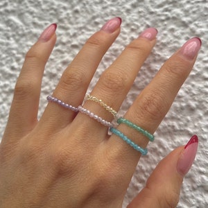 Può includere: Una mano mostra cinque anelli di perline in vari colori: lavanda, rosa pallido, giallo, azzurro e verde. Gli anelli sono delicati, con piccole perline infilate. Sfondo bianco strutturato.