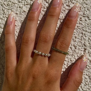 Può includere: Una mano con unghie lunghe e a forma di mandorla dipinte con uno smalto rosa chiaro e disegni di margherite bianche. La mano indossa due anelli, uno con piccole perle bianche e l'altro con perline verdi.
