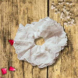 May include: White cotton hair scrunchie with a delicate lace trim. The scrunchie is lying on a wooden surface.