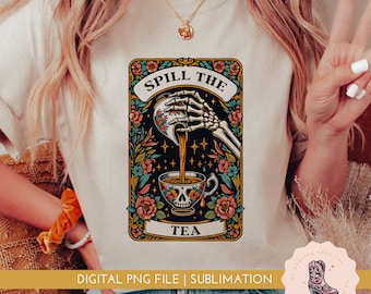 May include: A white t-shirt with a graphic design of a skeleton hand pouring tea from a skull-shaped teacup. The graphic is surrounded by floral elements and the text "Spill the Tea".