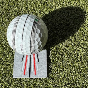May include: A white golf ball with black and gray markings sits on a gray alignment tool with red lines. The tool has the text "TaylorMade" printed on it.