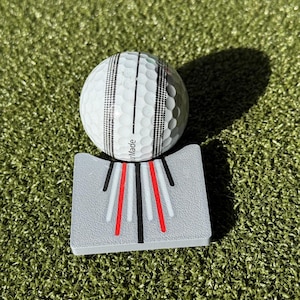 May include: A white golf ball with black stripes sits on a grey golf training aid with red and white stripes. The aid has the word "Made" printed on it.
