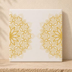 Op de afbeelding: Een vierkant canvas met een witte achtergrond en twee symmetrische, ingewikkelde gouden mandalapatronen die aan de linker- en rechterkant zijn gespiegeld. De ontwerpen bestaan uit herhalende bloemvormen, gebogen lijnen en geometrische bloemblaadjes in goud.