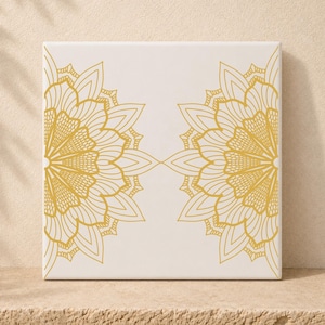 Könnte beinhalten: Eine quadratische weiße Keramikfliese mit zwei symmetrischen goldenen Mandala-Mustern. Jedes komplexe Design besteht aus geschichteten Blütenblättern und feinen Linien, die sich auf der linken und rechten Seite spiegeln und ein ausgewogenes Bild ergeben.
