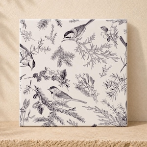 May include: A square ceramic tile featuring a black and white botanical illustration. The pattern displays small birds perched on evergreen branches, interspersed with clusters of berries and pine needles against a light cream background.
