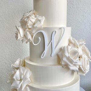 May include: A four-tier white wedding cake decorated with white roses and a white cursive letter 'W' on the third tier.