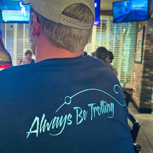 May include: A person wearing a blue t-shirt with the text "Always Be Trolling" in a light blue color. The person is wearing a gray mesh baseball cap.