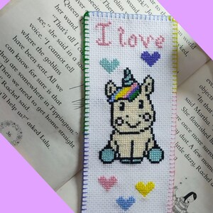 Könnte beinhalten: Ein weißer Kreuzstich-Lesezeichen mit einem bunten Einhorn-Design und dem Text "I love" in rosa Buchstaben. Das Einhorn hat eine Regenbogenmähne und einen Regenbogen-Schwanz, und es gibt vier Herzen in verschiedenen Farben um das Einhorn herum.