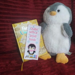 Pode incluir: Um close-up de um brinquedo de pelúcia de pinguim sentado ao lado de um livro com um marcador que diz "Chillin' with a good book". O marcador apresenta um pinguim usando uma echarpe rosa e segurando uma pilha de livros.