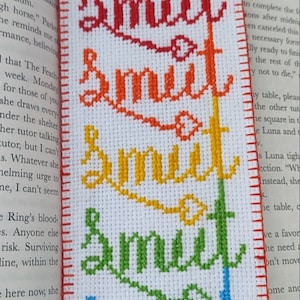 May include: A white bookmark with the word "Smut" embroidered in rainbow colors. The bookmark is made with a cross-stitch technique.