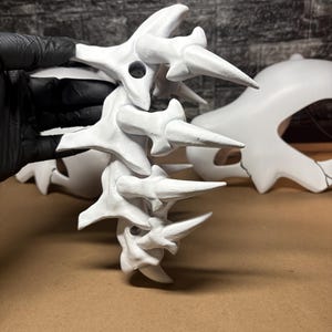 May include: A white sculpture, possibly a spine or vertebrae, with multiple segments and sharp, pointed projections. The piece is held by a gloved hand. Another, larger white sculpture is visible in the background.