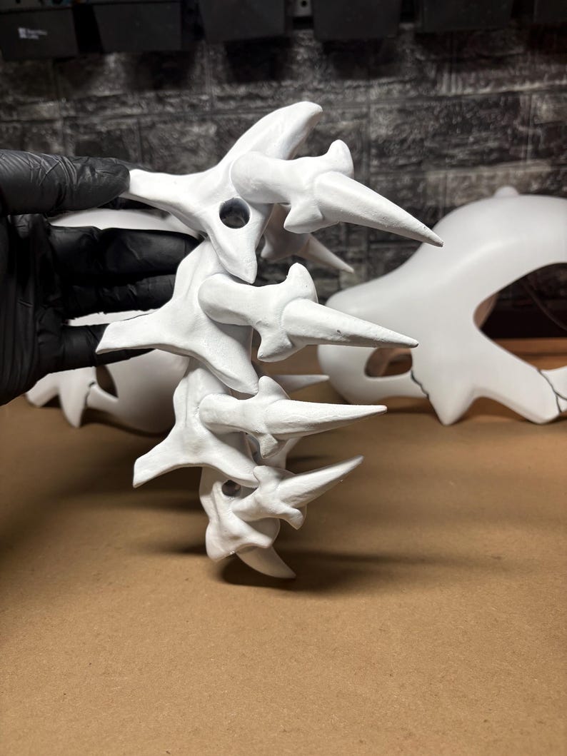 May include: A white sculpture, possibly a spine or vertebrae, with multiple segments and sharp, pointed projections. The piece is held by a gloved hand. Another, larger white sculpture is visible in the background.