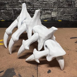 May include: A white, sculpted object resembling a skeletal claw or hand. It features multiple finger-like projections with sharp, pointed tips. The object is set against a textured, dark background.