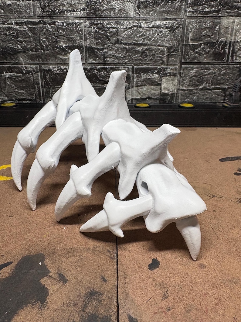 May include: A white, sculpted object resembling a skeletal claw or hand. It features multiple finger-like projections with sharp, pointed tips. The object is set against a textured, dark background.