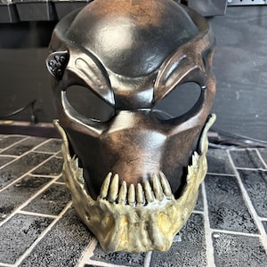 May include: A Predator mask with a brown and black paint scheme. The mask has a detailed jaw with sharp teeth painted in a gold color.