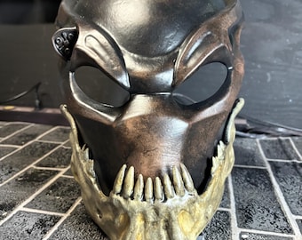 May include: A Predator mask with a brown and black paint scheme. The mask has a detailed jaw with sharp teeth painted in a gold color.