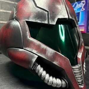 May include: A full-face helmet featuring a weathered red and black finish with a metallic sheen. The design includes a dark green tinted visor, a prominent angular brow, and a silver-coloured ribbed breathing apparatus attached to the lower chin area.