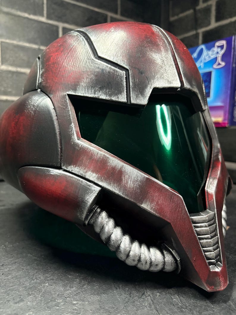 May include: A full-face helmet featuring a weathered red and black finish with a metallic sheen. The design includes a dark green tinted visor, a prominent angular brow, and a silver-coloured ribbed breathing apparatus attached to the lower chin area.
