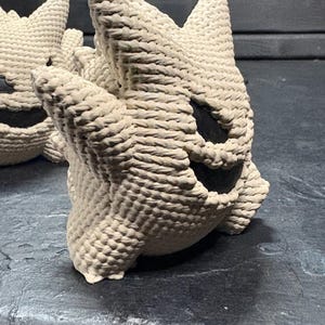 Pode incluir: Uma figura de croch&ecirc; na cor creme com cabe&ccedil;a e orelhas pontiagudas, apresentando uma boca larga e escura em forma de meia-lua e dois olhos escuros ovais. A textura &eacute; composta por pontos de fio grossos e vis&iacute;veis, criando uma forma arredondada.