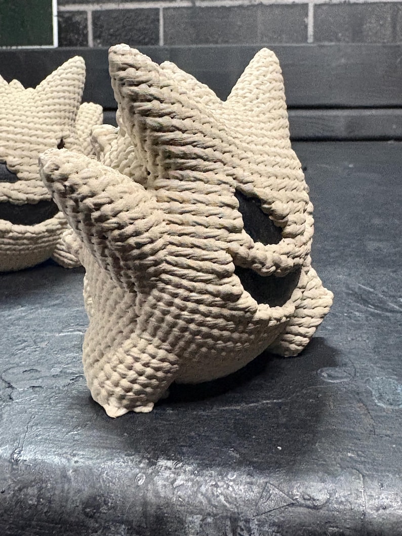 Pode incluir: Uma figura de croch&ecirc; na cor creme com cabe&ccedil;a e orelhas pontiagudas, apresentando uma boca larga e escura em forma de meia-lua e dois olhos escuros ovais. A textura &eacute; composta por pontos de fio grossos e vis&iacute;veis, criando uma forma arredondada.