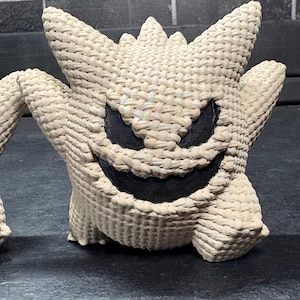 Pode incluir: Uma figura texturizada de cor creme, tecida, que se assemelha ao personagem Gengar com orelhas pontiagudas, um sorriso largo e &oacute;rbitas oculares escuras e profundas. O objeto apresenta uma constru&ccedil;&atilde;o densa tipo corda com um padr&atilde;o cruzado.