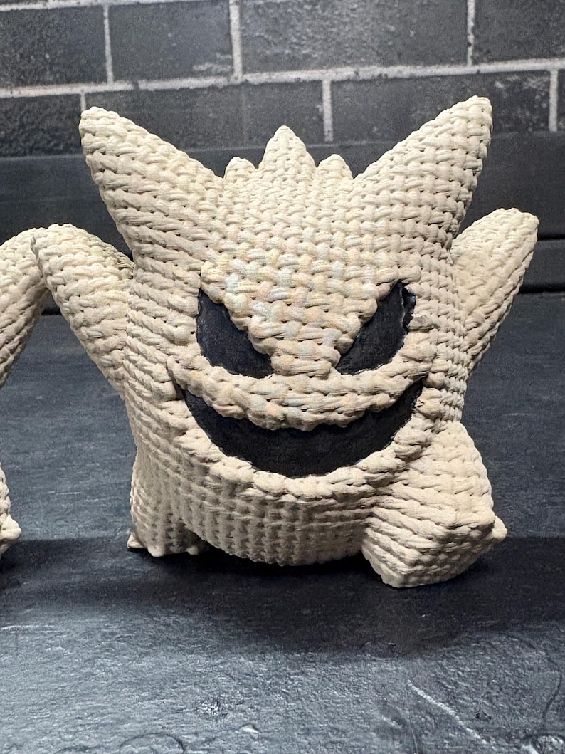 Pode incluir: Uma figura texturizada de cor creme, tecida, que se assemelha ao personagem Gengar com orelhas pontiagudas, um sorriso largo e &oacute;rbitas oculares escuras e profundas. O objeto apresenta uma constru&ccedil;&atilde;o densa tipo corda com um padr&atilde;o cruzado.