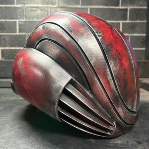 May include: A full-head helmet featuring a layered, curved design with a weathered finish. The surface displays a distressed red and metallic silver colour scheme with deep black accents. The side includes a series of horizontal vents or fins.