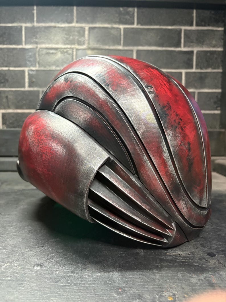May include: A full-head helmet featuring a layered, curved design with a weathered finish. The surface displays a distressed red and metallic silver colour scheme with deep black accents. The side includes a series of horizontal vents or fins.