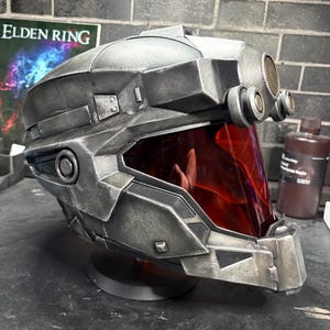 May include: A metallic gray sci-fi helmet featuring a weathered finish, a prominent red visor, and circular side attachments. The helmet has a structured, angular design with a protruding front section and a smooth, rounded top dome.