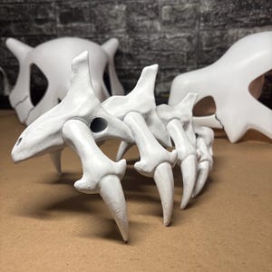 May include: White, 3D-printed Cubone skull and bone tail prop. The skull has a prominent jawline and eye sockets. The bone tail has a series of pointed, curved bone segments. The props are displayed on a brown surface.