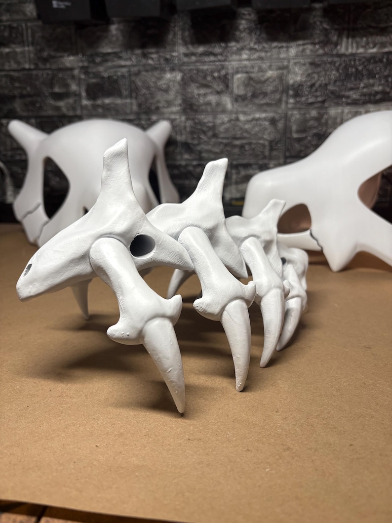 May include: White, 3D-printed Cubone skull and bone tail prop. The skull has a prominent jawline and eye sockets. The bone tail has a series of pointed, curved bone segments. The props are displayed on a brown surface.