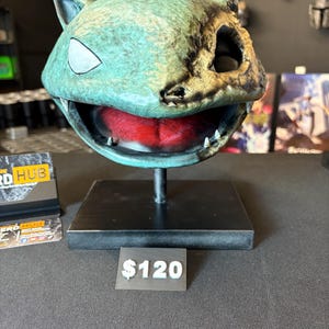 May include: A teal and weathered-looking creature head sculpture with a wide open mouth revealing a bright red interior, white pointed teeth, and a single visible white eye. The sculpture is mounted on a black rectangular base and stand.