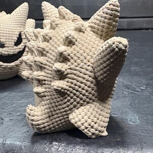Pode incluir: Uma figura de croch&ecirc; bege com corpo arredondado, m&uacute;ltiplos espinhos pontiagudos nas costas e membros curtos. A textura &eacute; criada com um padr&atilde;o de fio tecido denso. Uma segunda figura parcialmente vis&iacute;vel com rosto escuro est&aacute; posicionada ao lado.
