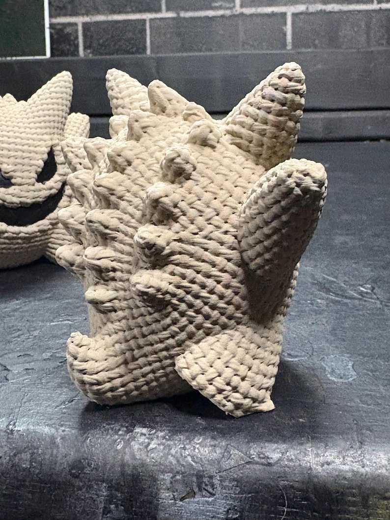 Pode incluir: Uma figura de croch&ecirc; bege com corpo arredondado, m&uacute;ltiplos espinhos pontiagudos nas costas e membros curtos. A textura &eacute; criada com um padr&atilde;o de fio tecido denso. Uma segunda figura parcialmente vis&iacute;vel com rosto escuro est&aacute; posicionada ao lado.