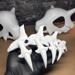 May include: A white claw prop with sharp, pointed tips, held by a gloved hand. Two white skull-shaped props are in the background. The props appear to be made of a hard material and are painted white.