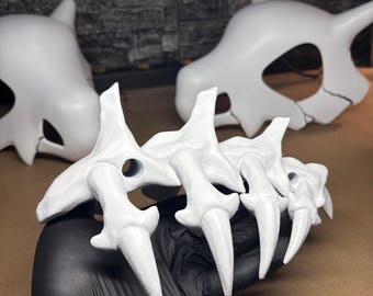 May include: White, claw-like prop with sharp, pointed tips, held by a gloved hand. Two additional white skull-shaped props are in the background. The props appear to be made of a hard material and are painted white.