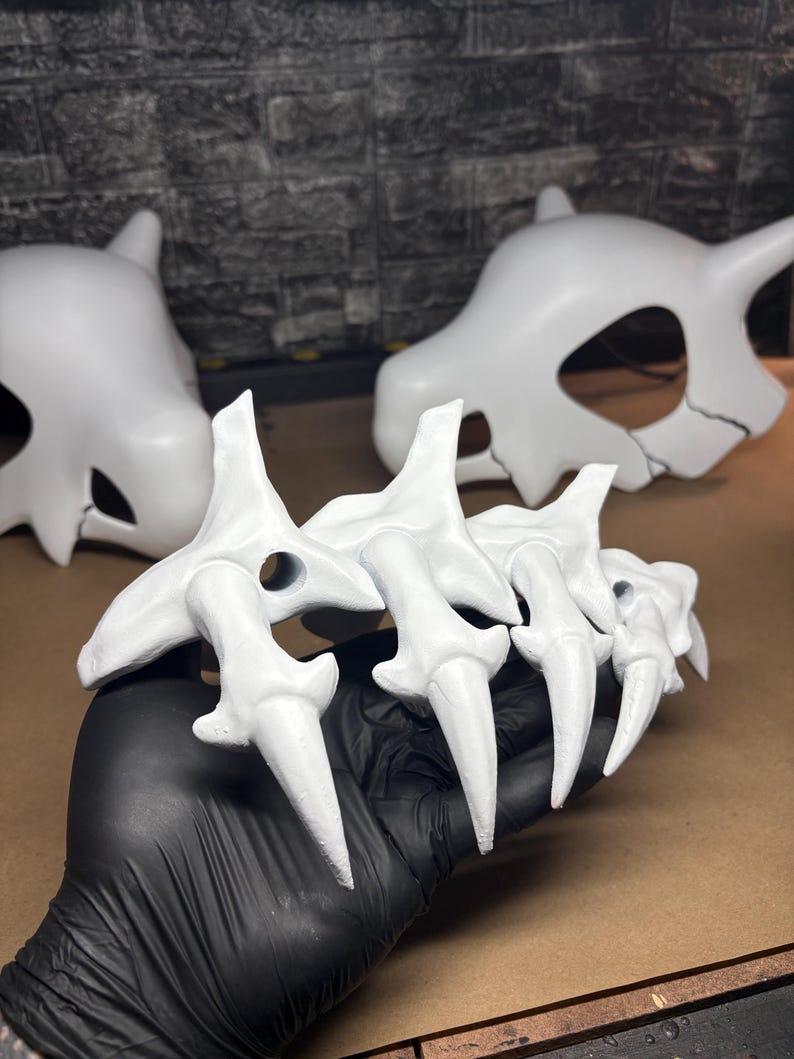 May include: A white claw prop with sharp, pointed tips, held by a gloved hand. Two white skull-shaped props are in the background. The props appear to be made of a hard material and are painted white.