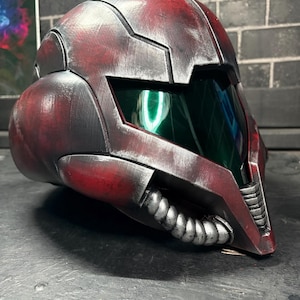 May include: A full-face helmet featuring a weathered red and black finish with a metallic sheen. The design includes a dark green tinted visor, segmented armour plating, and a silver-coloured ribbed breathing apparatus attached to the lower front section.