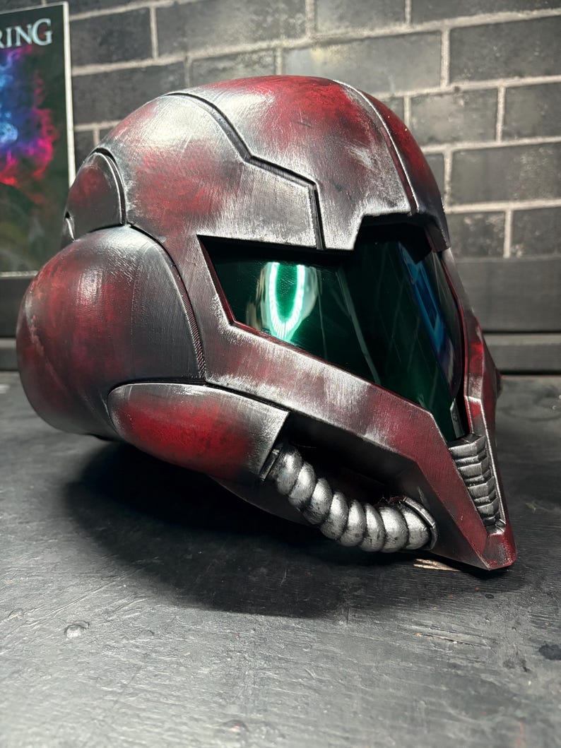 May include: A full-face helmet featuring a weathered red and black finish with a metallic sheen. The design includes a dark green tinted visor, segmented armour plating, and a silver-coloured ribbed breathing apparatus attached to the lower front section.