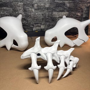 May include: Three white, stylized bone structures are displayed on a brown surface. Two skull-shaped objects are in the background, and a spine-like structure is in the foreground. The objects appear to be props or models.