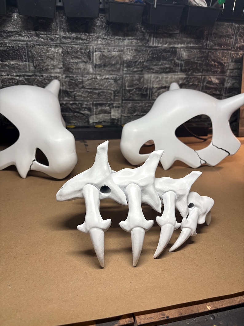 May include: Three white, stylized bone structures are displayed on a brown surface. Two skull-shaped objects are in the background, and a spine-like structure is in the foreground. The objects appear to be props or models.