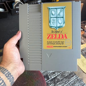 May include: A gray Nintendo Entertainment System game cartridge for "The Legend of Zelda." The label features a gold background with a crest design and the game's title in red and white. Includes invaluable maps and strategic playing tips.
