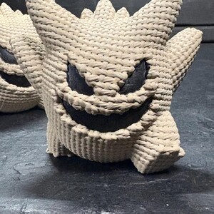 Pode incluir: Uma figura de croch&ecirc; bege com corpo arredondado, orelhas pontiagudas e espinhos na cabe&ccedil;a. A figura apresenta um sorriso largo e amea&ccedil;ador e olhos triangulares afiados preenchidos com material preto, contrastando com o fio texturizado.
