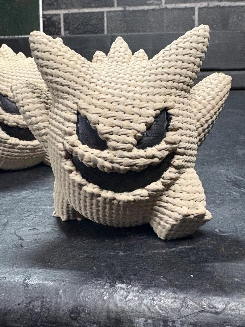 Pode incluir: Uma figura de croch&ecirc; bege com corpo arredondado, orelhas pontiagudas e espinhos na cabe&ccedil;a. A figura apresenta um sorriso largo e amea&ccedil;ador e olhos triangulares afiados preenchidos com material preto, contrastando com o fio texturizado.