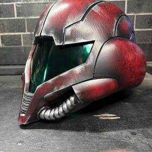 May include: A full-coverage helmet featuring a weathered red and black finish with a metallic sheen. The design includes a dark green tinted visor, a prominent central ridge, and a silver-coloured ribbed tube detail along the lower jaw area.