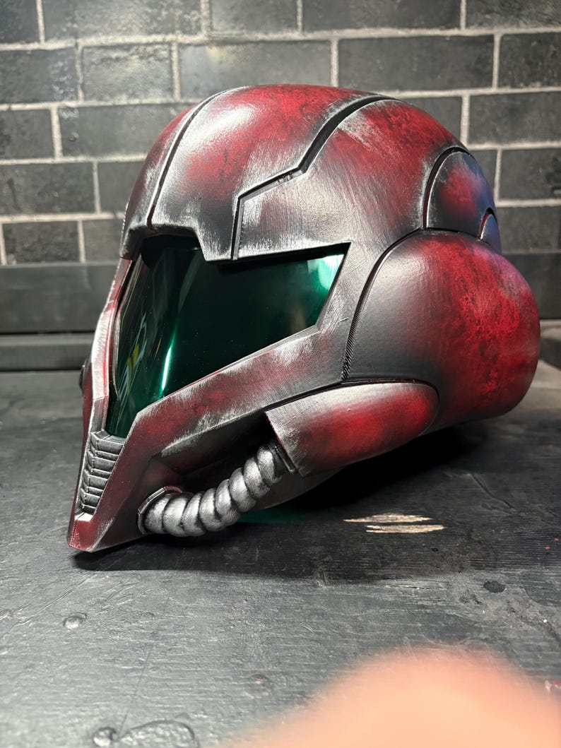 May include: A full-coverage helmet featuring a weathered red and black finish with a metallic sheen. The design includes a dark green tinted visor, a prominent central ridge, and a silver-coloured ribbed tube detail along the lower jaw area.