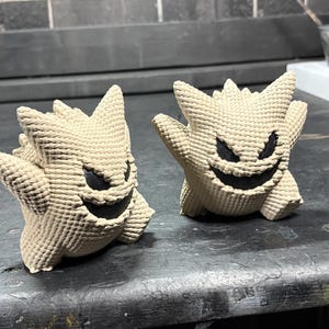 Pode incluir: Duas figuras bege texturizadas com a forma da personagem Gengar, com orelhas pontiagudas, costas espinhosas e bocas largas, pretas e sorridentes com olhos triangulares afiados. As figuras t&ecirc;m um padr&atilde;o de superf&iacute;cie tecido distinto.