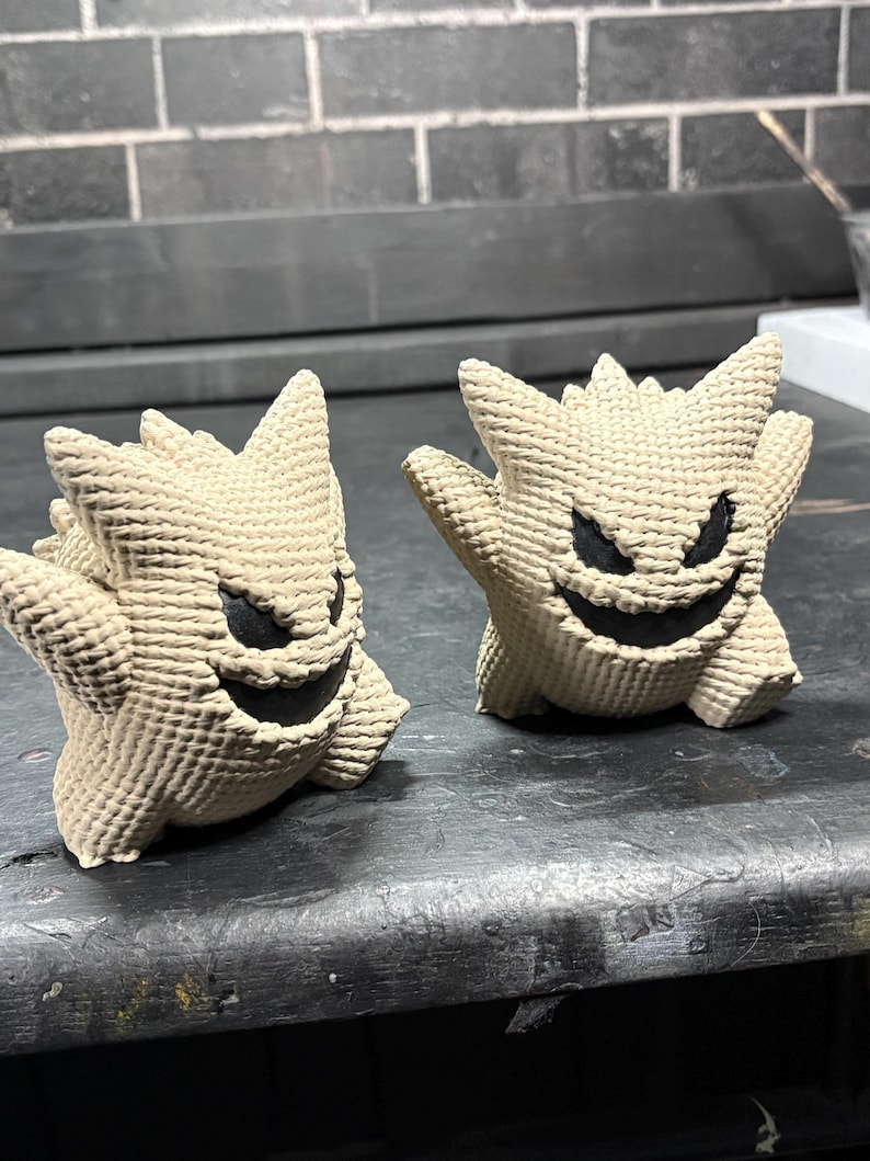 Pode incluir: Duas figuras bege texturizadas com a forma da personagem Gengar, com orelhas pontiagudas, costas espinhosas e bocas largas, pretas e sorridentes com olhos triangulares afiados. As figuras t&ecirc;m um padr&atilde;o de superf&iacute;cie tecido distinto.